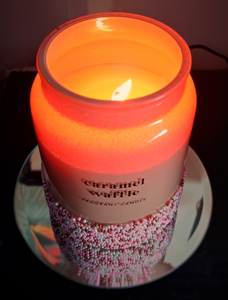 A scented Caramel Waffle candle glowing brightly in its jar covered with Hundreds And Thousands.