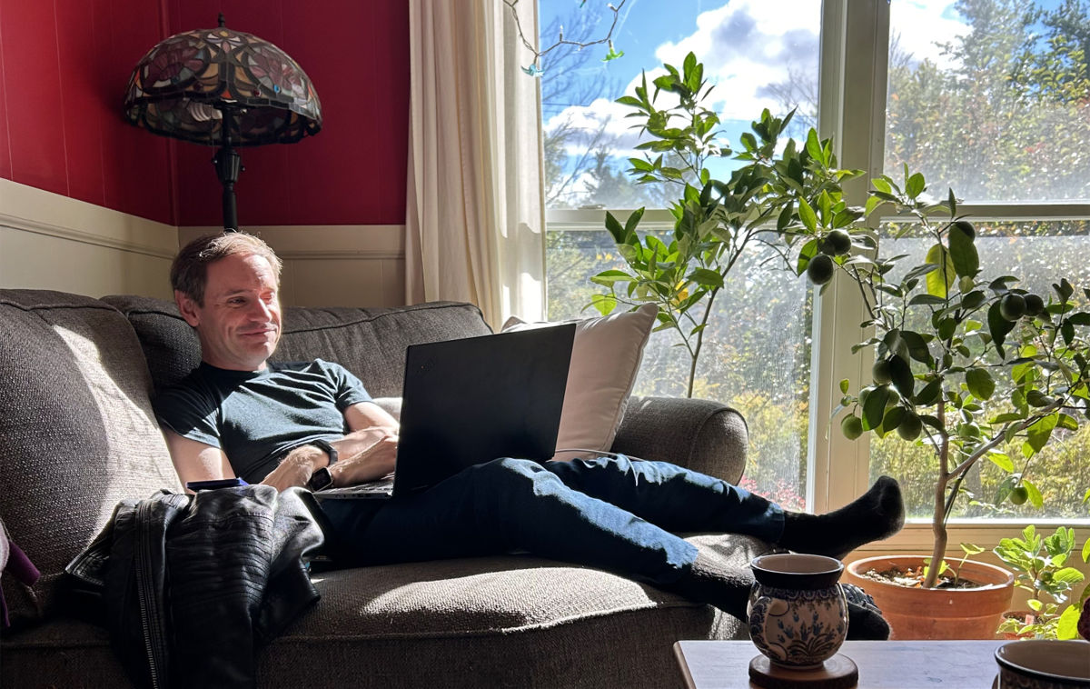Kohan Ikin smiling and relaxed while using his laptop computer, in a colorful sunny room surrounded by plants in Ithaca.