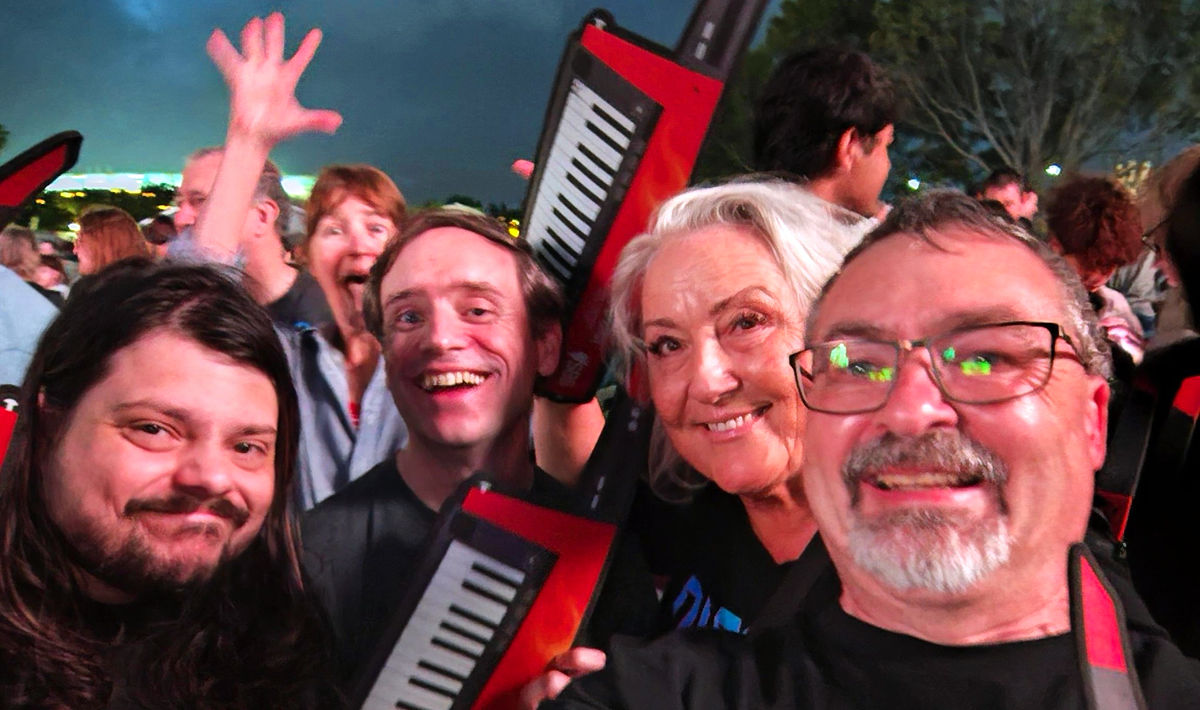 Michael Romano, Kohan Ikin and Kate Hansen cheering while watching Voyager perform at the WA Day 2025 concert on 22 November 2025. They are holding signs that resemble Voyager's red Korg keytar synthesizer.